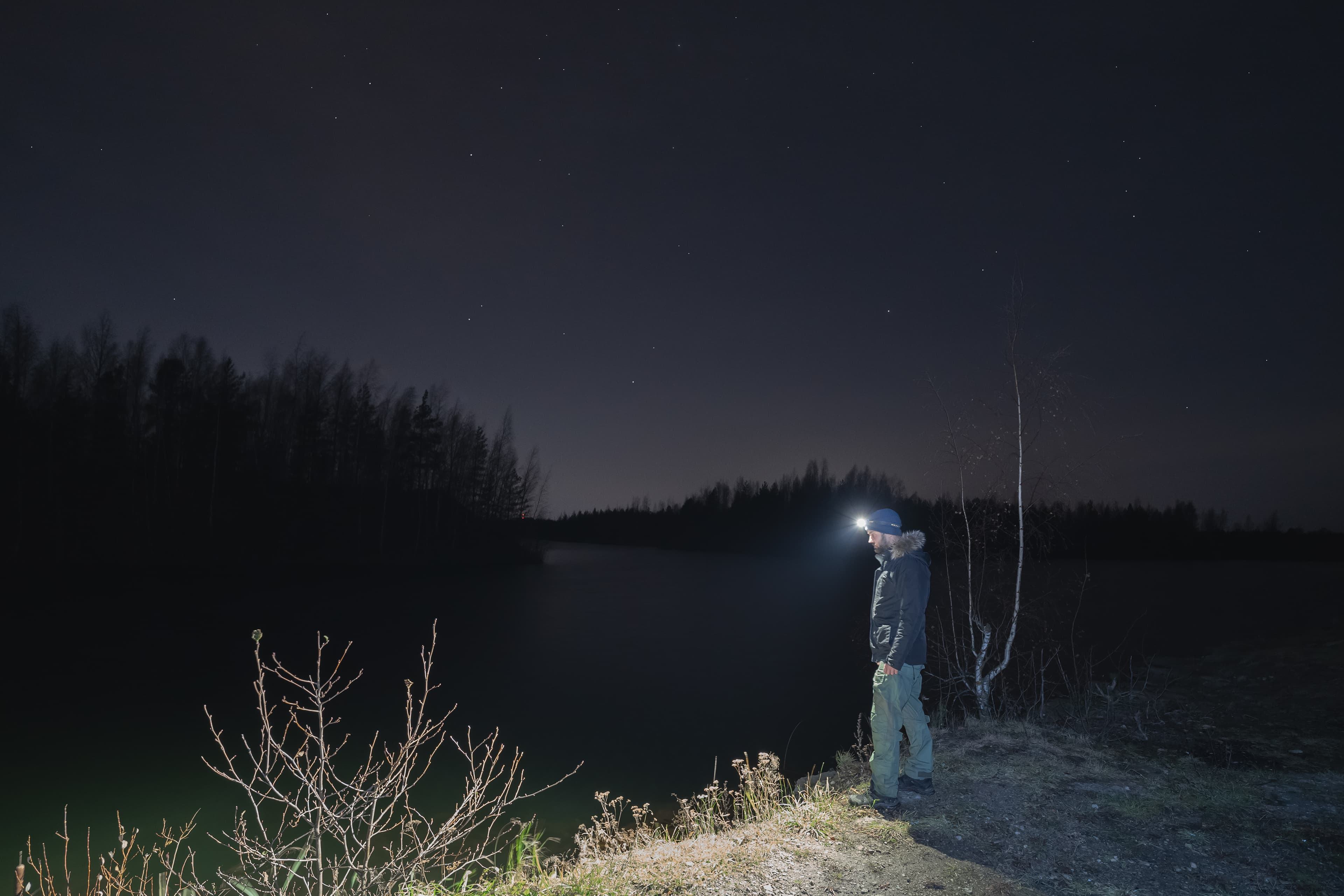 A man wearing a rechargeable headlamp lights the darkness beside a lake at night under a starry sky
